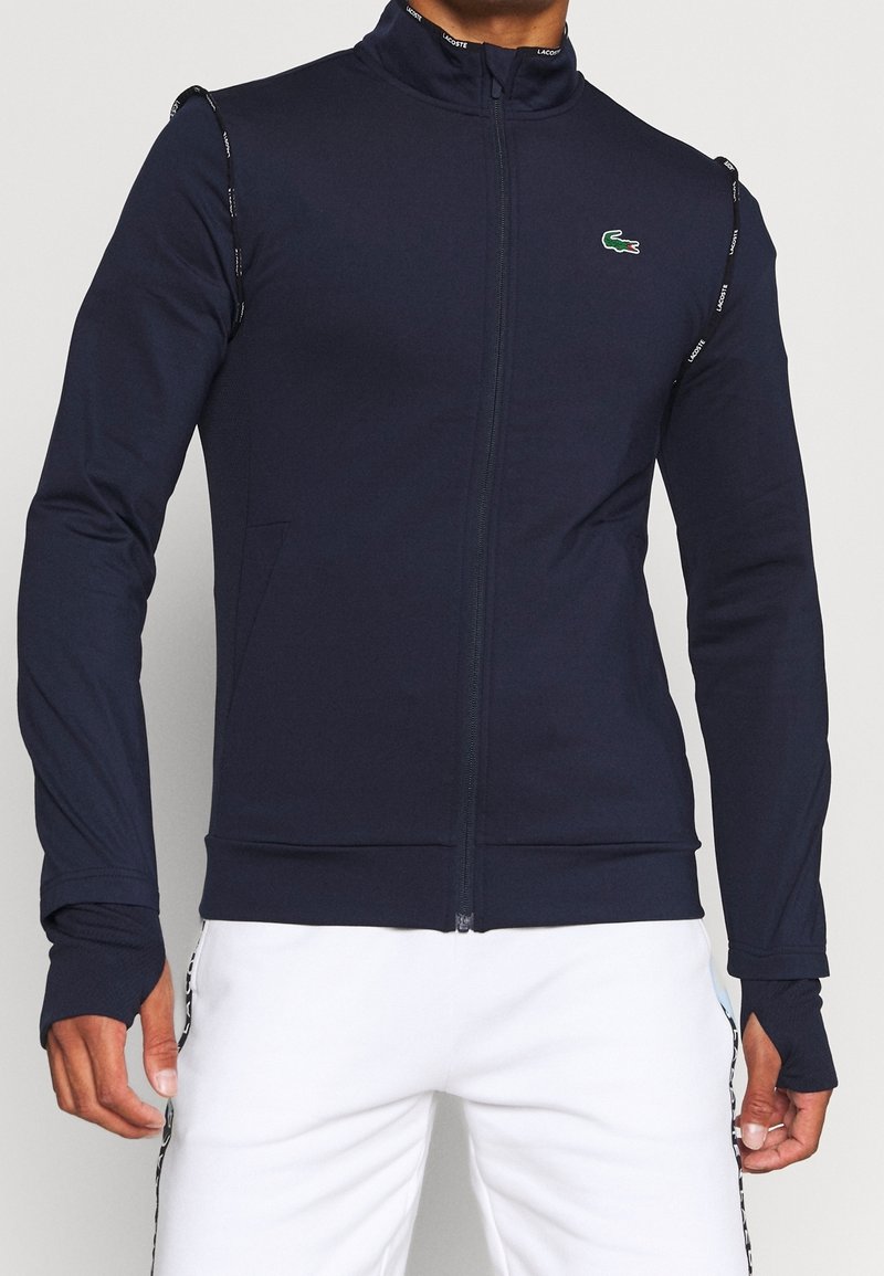 Marin zip-up jacka i stretchig tyg med hög krage, tumhål och Lacoste-logotyp på bröstet. Har sidofickor och kontrasterande kant.