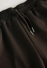 Braune Jogginghose mit elastischem Bund, ausgestattet mit einem Kordelzug und silbernem Zubehör. Der Stoff hat eine glatte, strukturierte Oberfläche.