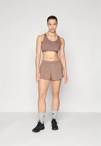 Gaiši brūns sporta krūšturis un atbilstoši šorti no elpojoša auduma. Melni sporta apavi, pelēkas zeķes un vienkāršs dizains ar minimālu zīmola apzīmējumu.