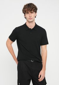 Νέος άνδρας με σγουρά μαλλιά, που φοράει μαύρο πόλο με κοντά μανίκια και μαύρο παντελόνι, ποζάρει μπροστά σε λευκό φόντο.