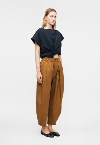 Γυναίκα όρθια, φοράει χοντρά καφέ ψηλόμεσα παντελόνια, μαύρο κοντομάνικο τοπ με κόμπο, και μαύρα επίπεδα παπούτσια, με ίσια ανοιχτόχρωμα μαλλιά.