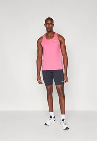 Roza brezrokavnik s logotipom, v kombinaciji s temno sivimi tesno prilegajočimi kratkimi hlačami. Bele športne superge. Nevtralna podlaga.