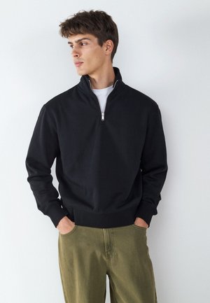 Jonge man met een zwarte pullover met korte rits en olijfgroene broek, handen in de zakken, kijkt zijwaarts tegen een witte achtergrond.
