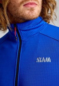 Primo piano del volto inferiore e del collo di una persona che indossa una giacca zip-up blu con texture, dotata di una zip rossa e un logo ricamato bianco.