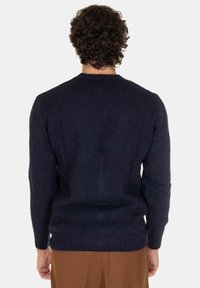 Uomo con capelli ricci che indossa un maglione blu scuro e pantaloni marroni, in piedi con la schiena rivolta verso uno sfondo chiaro e semplice.