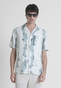 Camicia a maniche corte in tessuto leggero con un motivo astratto verde, dotata di collo a punte e chiusura con bottoni. Indossata con occhiali da sole.
