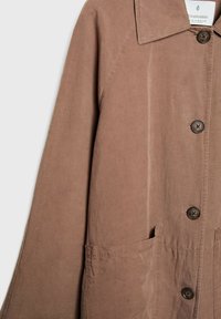 Bruine katoenen jas met een puntige kraag, knoopsluiting, twee zijzakken en zichtbare stikdetails. Geweven stof met een zachte afwerking.