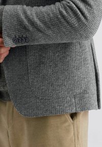 Grijze geruite jas van een gestructureerde stof. Heeft een notch lapel, zijzakken en vier knopen op de manchetten in een donkere tint.