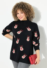 Schwarzer Strickpullover mit hohem Kragen, versehen mit mehrfarbigen Rentiermotiven. Das Modell hält eine rote gesteppte Clutch. Graue Jeans runden den Look ab.