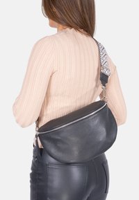 Schwarze Leder-Umhängetasche mit curved Form, ausgestattet mit einem silbernen Reißverschluss und einem strukturierten Gurt mit geometrischem Muster.