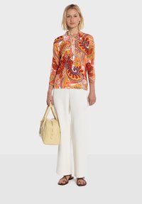 Haut à manches longues à imprimé paisley multicolore, tissu léger, associé à un pantalon large blanc, portant un sac à main jaune, chaussures marron.