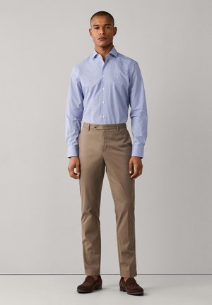 Man staat, draagt een lichtblauw overhemd met knopen, beige broek en donkerbruine loafers tegen een effen lichte achtergrond.