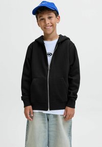 Crna zip-up hoodie s prednjim džepovima, s kapuljačom na povlačenje, nosi se preko bijele majice i uparen je s svijetloplavim trapericama.