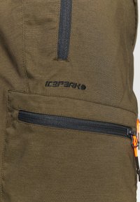 Lähikuva oliivinvihreästä kankaasta, jossa on kaksi mustaa vetoketjua ja musta Icepeak-logo brodeerattuna.