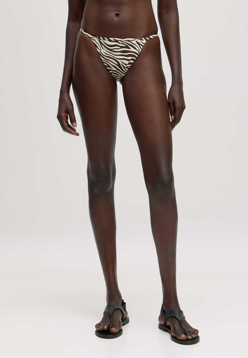 Zebra-gepatroneerde bikini onderkant in crème en bruine tinten, low-rise ontwerp, dunne zijbandjes, gecombineerd met zwarte minimalistische sandalen.