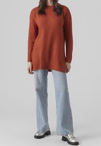 Femme portant un long pull orange brûlé, un jean large bleu clair et des mocassins à semelle épaisse argentés, debout devant un fond gris.