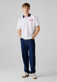 Hvid polo-shirt med en krave i rød og marineblå, prydet med "Red Bull RACING" broderet i rød. Båret med mørkeblå jeans og hvide sneakers.