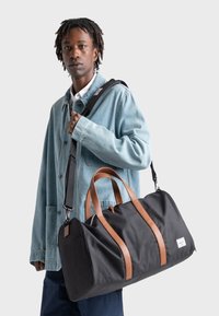 Sort duffeltaske med brune læderhåndtag og detaljer, lavet af tekstureret materiale, med en skulderrem og rektangulær form.