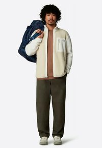 Mees, kes kannab beežist tõmblukuga fliisi, pruunist särgist, tumedatest pükstest ja valgetest tossudest, hoiab sinist spordikotti õlal, seistes valge tausta vastu.