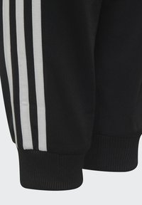 Pantalons de jogging en tissu noir avec trois rayures blanches verticales. Caractéristiques des poignets côtelés et un matériau texturé pour le confort.