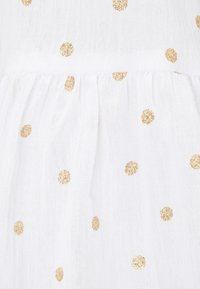 Tejido blanco con una superficie texturada y translúcida que presenta lunares dorados espaciados uniformemente, creando una apariencia delicada y ligera.
