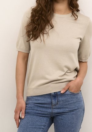 Pull en maille beige clair à manches courtes, col rond et ourlet côtelé ; associé à un jean denim bleu. Tissu texturé avec un éclat subtil.