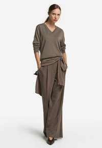 Kvinna står med händerna i fickorna, iförd en lös brun V-ringad tröja, vida byxor, en knuten skjorta runt midjan och mörka spetsiga skor.