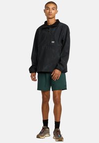 Schwarzer Halbzip-Pullover mit strukturiertem Muster, Brusttasche, kombiniert mit grünen Shorts und grauen Schuhen, stehend vor einem einfarbigen Hintergrund.