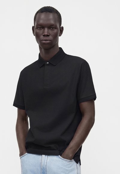 Homme portant un polo noir texturé et un jean bleu clair, debout les mains dans les poches devant un fond clair uni.