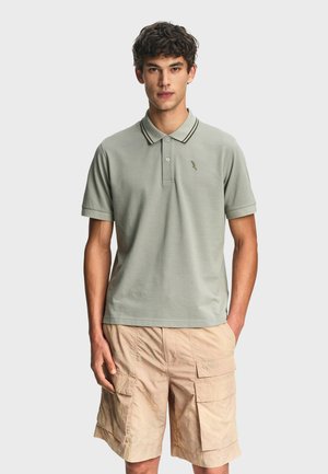 Uomo che indossa una polo a maniche corte verde chiaro e pantaloncini cargo beige, in piedi con le mani in tasca davanti a uno sfondo semplice.