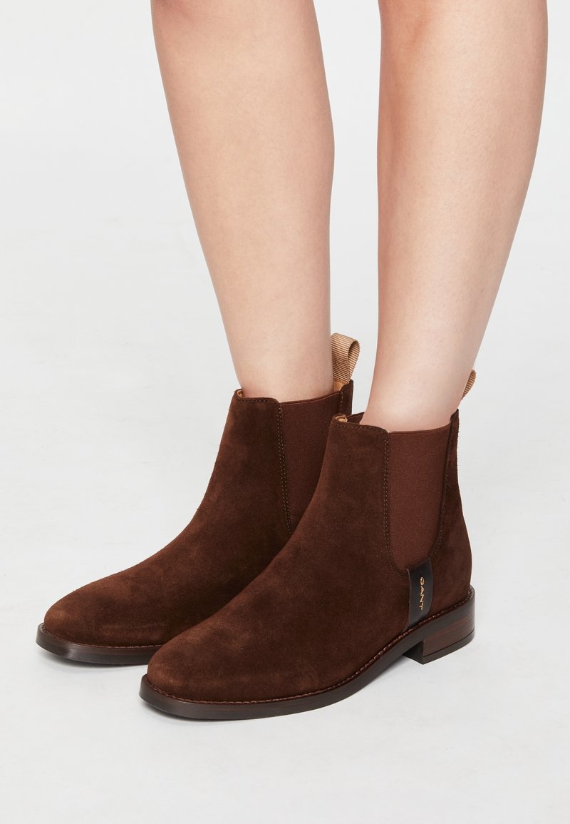 Bottines en daim marron à petit talon, avec panneaux latéraux élastiques et languettes, portées sur jambes nues sur un fond neutre.