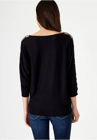 Svart stickad topp med lös passform, trekvartslånga ärmar och dekorativa knappdetaljer på axlarna. Ribbad fåll och mjuk textur.