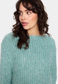 Šviesiai mėlynos žalios spalvos megztinis su laisvu siluetu ir storu, tekstūruotu audiniu. Turi apvalų iškirpimą ir nuleistas pečiais.