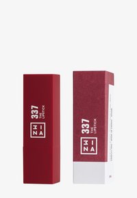 Röd lipstiftube med matt yta, försedd med vit text på framsidan. Följd av en matchande kartongbox med minimalistisk design.