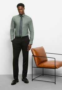 Grünes langärmeliges Hemd kombiniert mit schwarzer Hose und glänzenden schwarzen Schuhen; dazu eine gestreifte Krawatte. Im Hintergrund steht ein brauner Ledersessel.