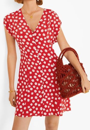 Femme portant une robe rouge fleurie avec des fleurs blanches, tenant un sac tressé rouge, avec des bijoux en or au poignet et au cou.