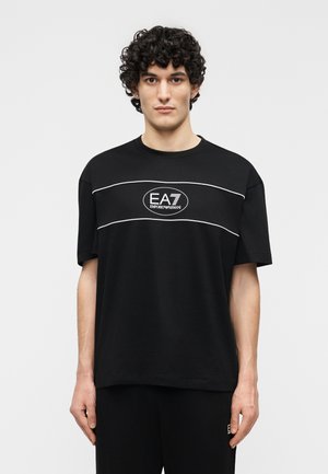 Młody mężczyzna o kręconych włosach, ubrany w czarny T-shirt EA7 Emporio Armani z białym obszyciem i logo na piersi, stojący na białym tle.
