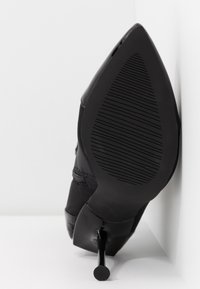 Zapato de tacón alto negro con puntera afilada y diseño elegante. Suela de goma texturizada con ranuras verticales y un delgado tacón de aguja.