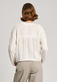 Weißer Strickpullover mit strukturiertem Muster, der breite Ärmel und ein kurzes Design aufweist. Die Rückseite zeigt ein offenes Strickdetail.