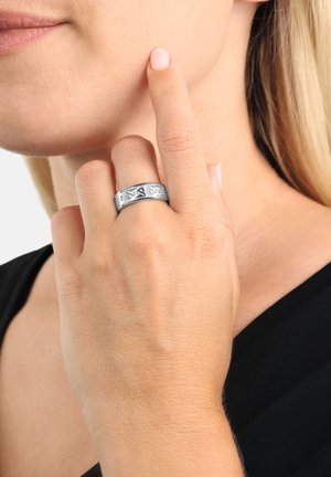 Silberner Ring mit kompliziertem eingraviertem Design, an einer Hand getragen, das seine glatte Textur und polierte Oberfläche vor einem dunklen Oberteil hervorhebt.