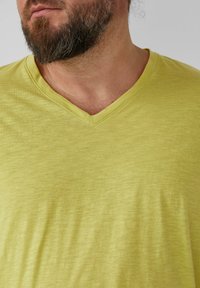 T-shirt z dekoltem w serek, krótkim rękawem, wykonany z lekkiego materiału w jasnym, limonkowym kolorze. Materiał wydaje się miękki, ma delikatną teksturę i lekki połysk.