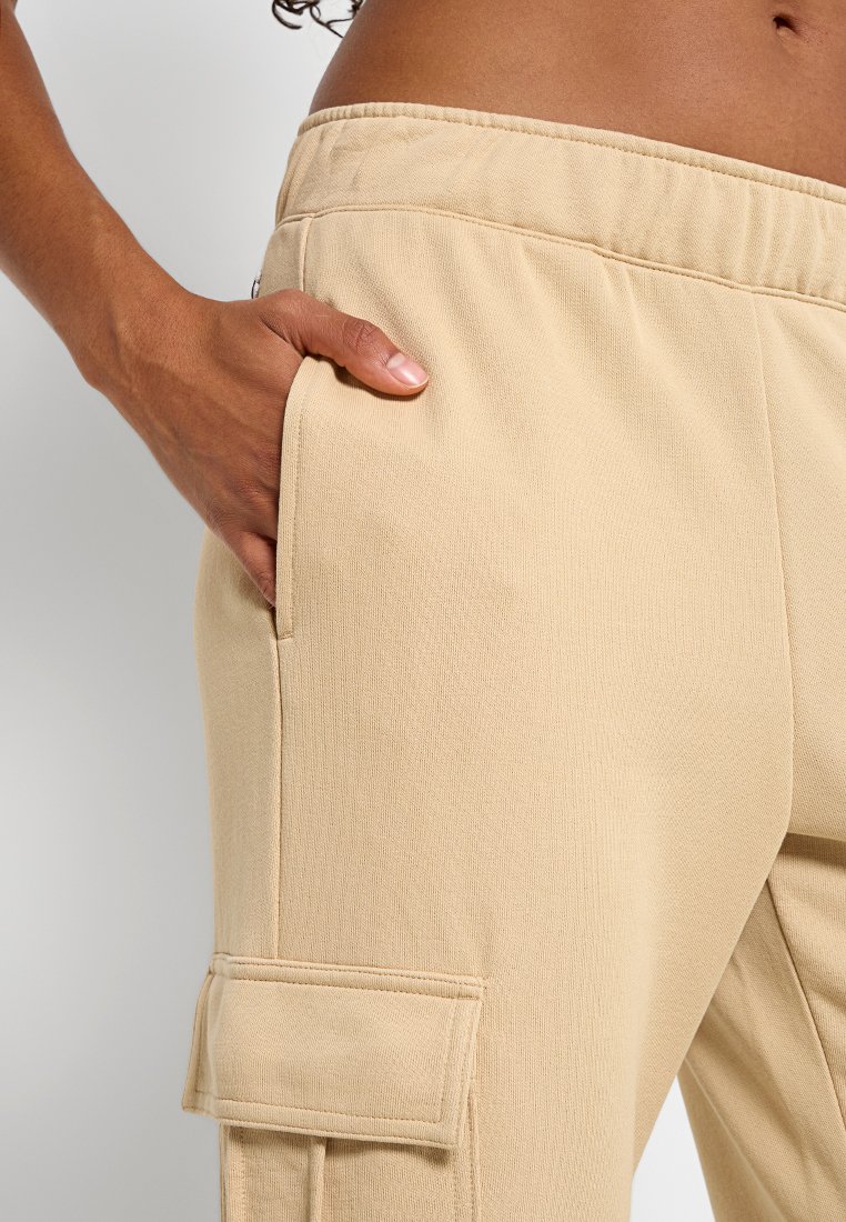 Le pantalon cargo beige présente une texture lisse, une taille élastique et une poche latérale avec un rabat, mettant en avant un design moderne et fonctionnel.