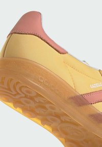 Zapatilla amarilla con un talón de cuero rosa y rayas rosas. Presenta una suela translúcida texturizada con patrones circulares y una marca.