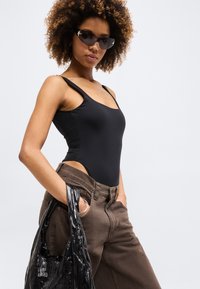 Γυναίκα που φοράει μαύρο τοπάκι, καφέ παντελόνι και σκούρα γυαλιά ηλίου κρατώντας μια μαύρη γυαλιστερή τσάντα με το ένα χέρι στην τσέπη, σε λευκό φόντο.