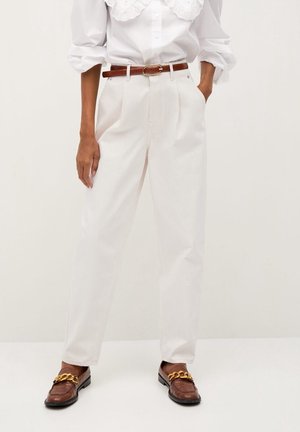 Femme portant un pantalon plissé blanc taille haute avec ceinture marron et mocassins marron avec détail chaîne dorée, mains dans les poches.