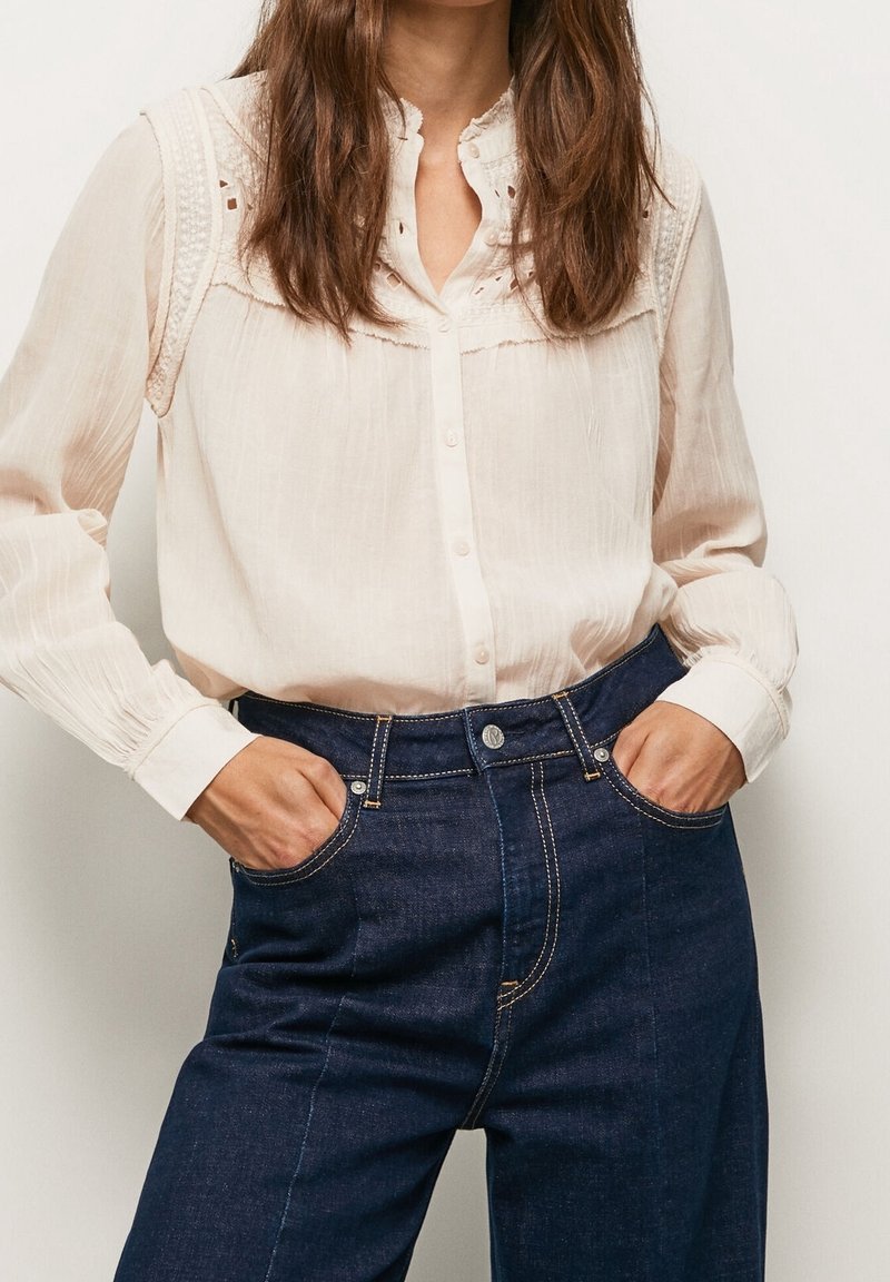 Camicia crema con bottoni, decorata con delicati ricami e colletto alto, abbinata a jeans in denim scuro a vita alta con cuciture a contrasto.