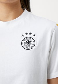 Λευκό βαμβακερό μπλουζάκι με κοντό μανίκι που διαθέτει ένα κυκλικό μαύρο και άσπρο κεντημένο λογότυπο με πέντε αστέρια από πάνω. Κοντά μανίκια με κίτρινες λεπτομέρειες.