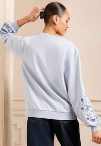 Svetlo modra jopica z rožnatim vezenjem na levem rokavu, z ribastimi manžetami in sproščenim krojem. Nosi se z temno jeanskimi hlačami.