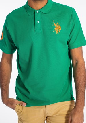 Mann trägt grünes Poloshirt mit orangefarbenem Pferd- und Reiter-Logo, beige Hose und Hand in der Tasche vor weißem Hintergrund.