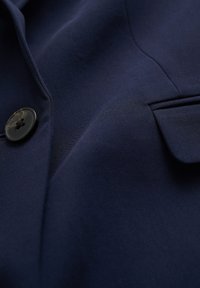 Námořnická modrá bunda s hladkou tkaninou, jedním černým knoflíkem a přizpůsobenou kapsou na levé straně.